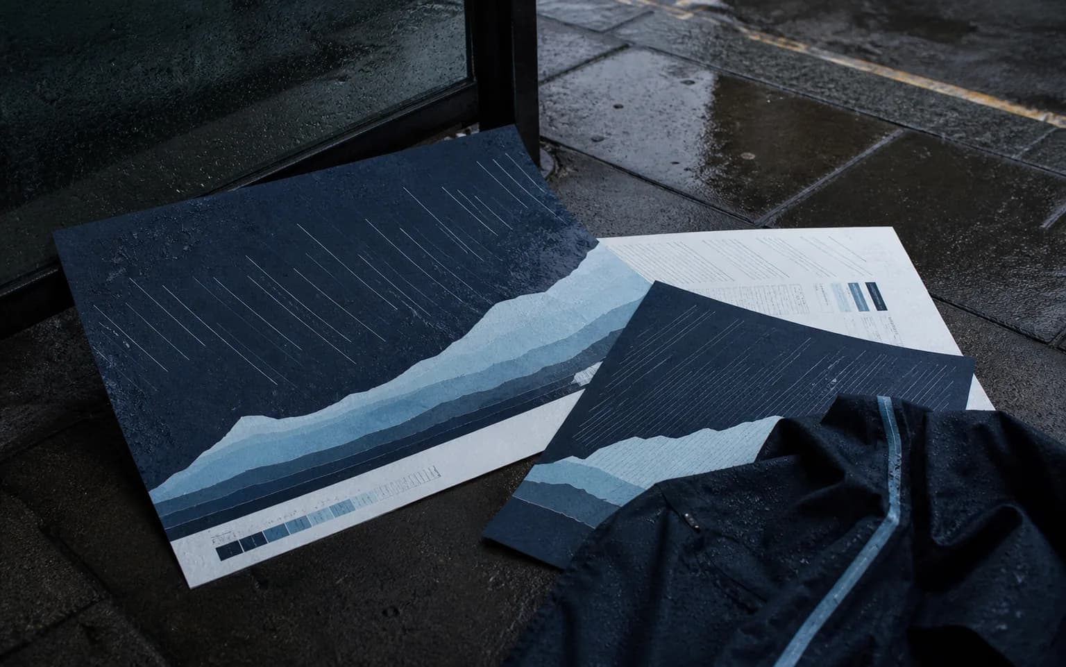 Rain-soaked Northfell campaign proofs and a dark waterproof garment arranged beside a wet bus shelter.